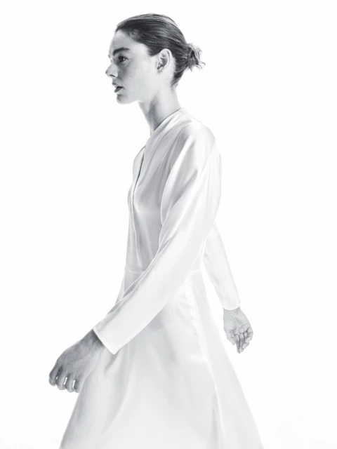 Monochromatic full shot of a slender adult woman walking in a dress with long sleeves against a bright white backdrop.