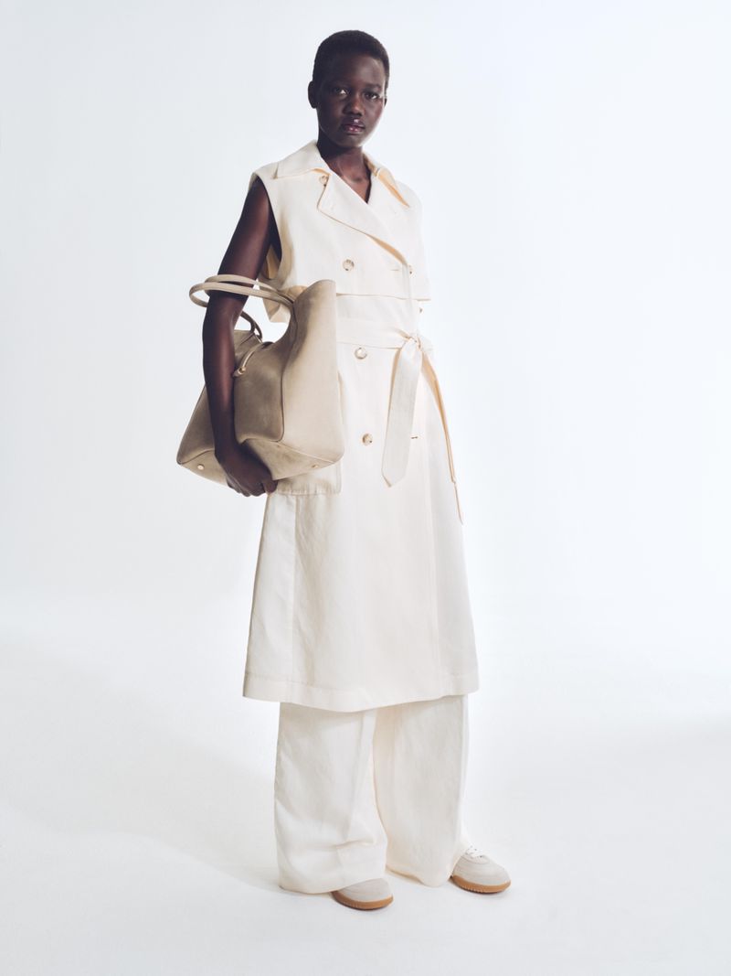 Full-length shot of an African woman in white vest and pants, holding a beige bag, in a clean studio fashion shot on a white background.