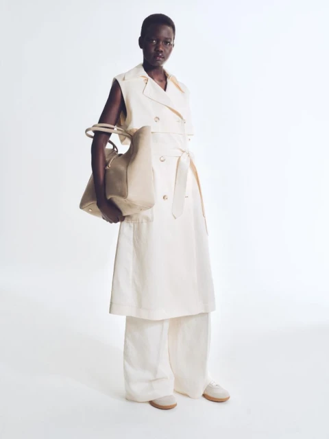 Full-length shot of an African woman in white vest and pants, holding a beige bag, in a clean studio fashion shot on a white background.