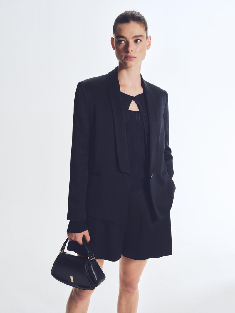 A woman in a black blazer and shorts poses with a black handbag against a white background in a fashion-forward studio setting.