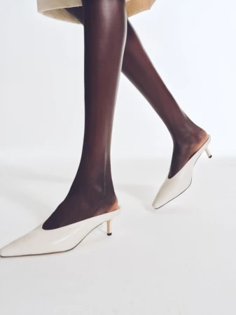 An African-American woman models a pair of beige, high-heeled, pointed-toe mules while standing against a neutral background in a studio.