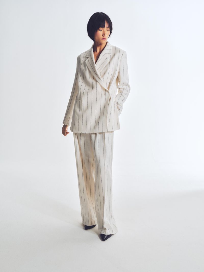 An Asian woman is full-length in a photo studio, wearing a cream-colored, pinstripe, double-breasted suit with wide-legged trousers.