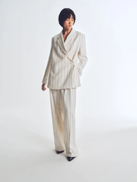 An Asian woman is full-length in a photo studio, wearing a cream-colored, pinstripe, double-breasted suit with wide-legged trousers.