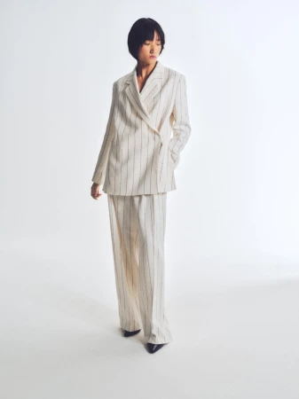 An Asian woman is full-length in a photo studio, wearing a cream-colored, pinstripe, double-breasted suit with wide-legged trousers.