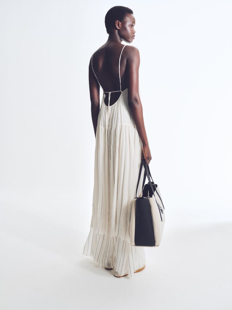 Back view of a Black female model in a flowing white dress holding a two-tone leather tote bag against a stark white background.