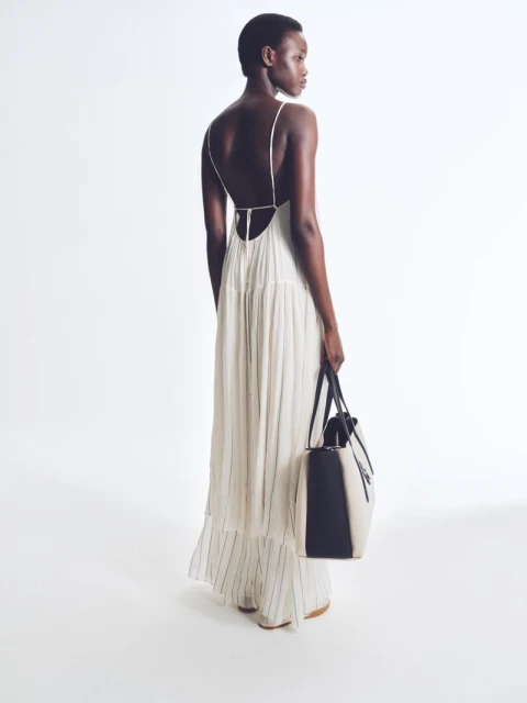 Back view of a Black female model in a flowing white dress holding a two-tone leather tote bag against a stark white background.