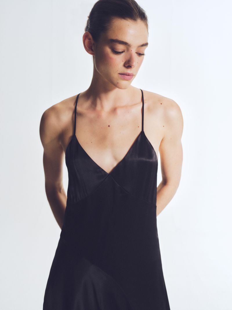 A woman with dark hair and fair skin wears a sleek black slip dress in a studio setting with soft, diffused lighting.