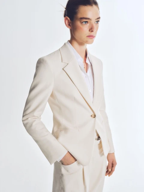 A woman models a stylish ivory blazer and matching pants in a bright studio. The blazer is single-button, paired with a white shirt.