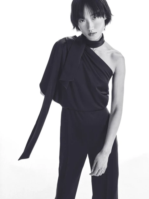 Elegant black and white fashion portrait of a woman in a draped, one-shoulder jumpsuit with tied neck detail, posing on a white background.
