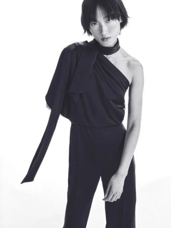 Elegant black and white fashion portrait of a woman in a draped, one-shoulder jumpsuit with tied neck detail, posing on a white background.