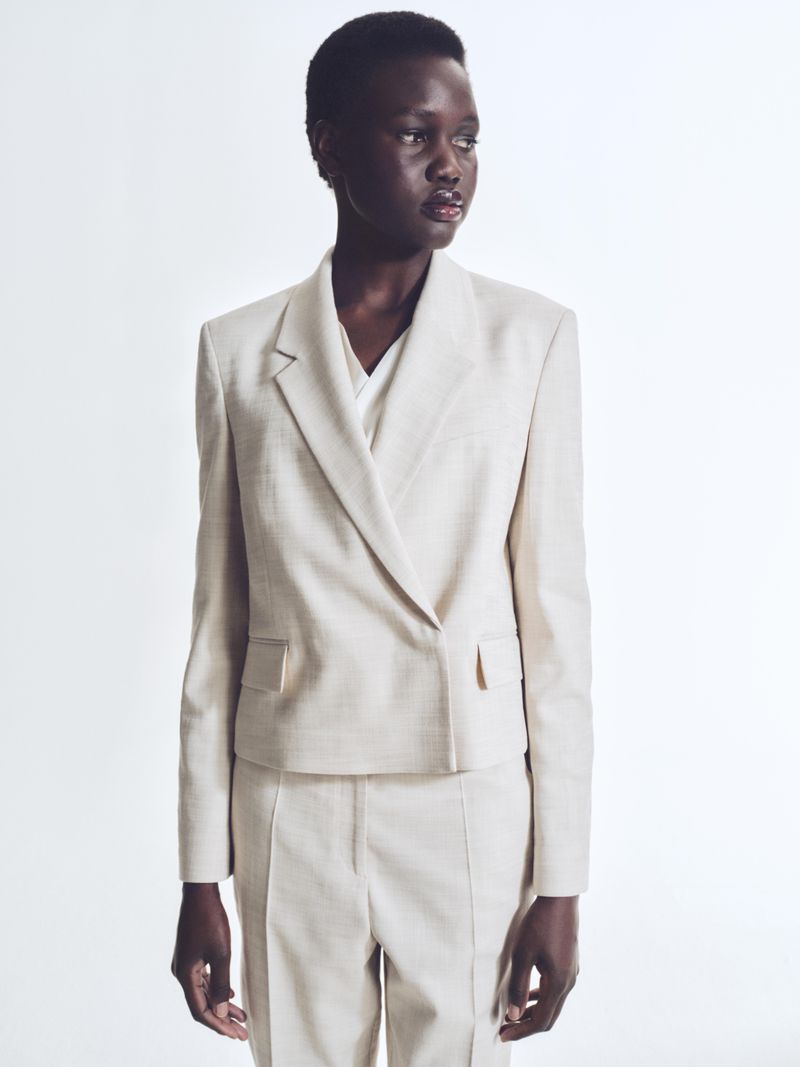 Adult woman in beige suit poses in a studio. She is wearing a double-breasted blazer and matching pants, standing against a white background.