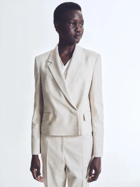Adult woman in beige suit poses in a studio. She is wearing a double-breasted blazer and matching pants, standing against a white background.