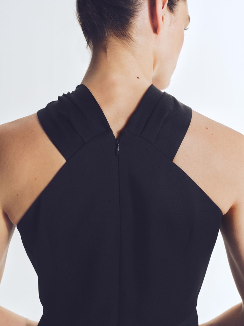 Back view of a woman with her hair up wearing a black criss-cross halter dress against a plain white background in an indoor setting.