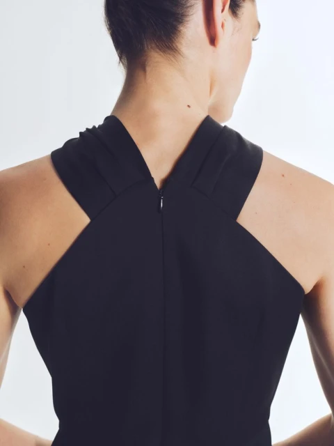 Back view of a woman with her hair up wearing a black criss-cross halter dress against a plain white background in an indoor setting.