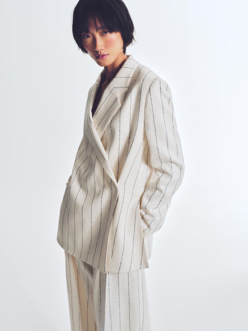 A slender Asian woman with short black hair poses in a cream-colored, pinstripe blazer and matching pants on a white backdrop.