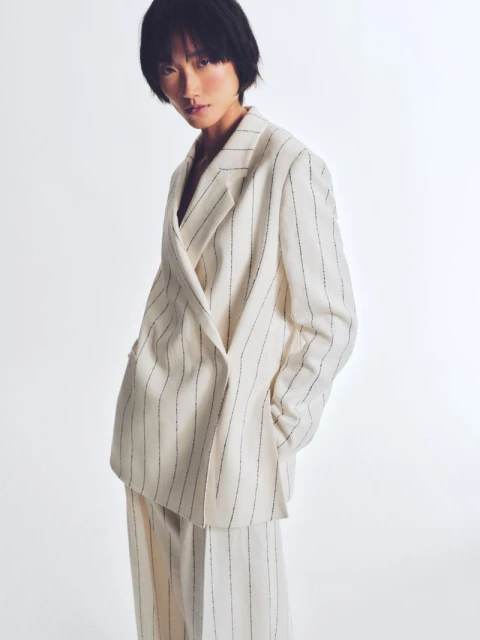 A slender Asian woman with short black hair poses in a cream-colored, pinstripe blazer and matching pants on a white backdrop.