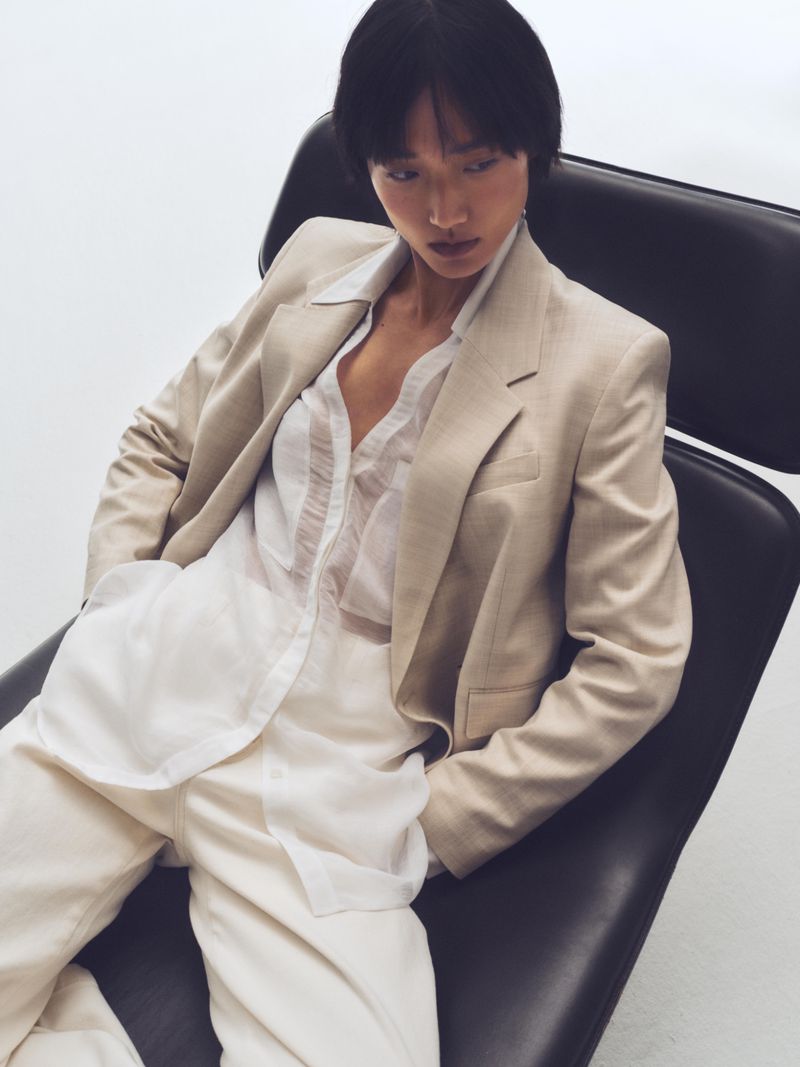 Woman in blazer and wide-leg pants sits in black modern chair against a neutral background in a fashion editorial-style portrait.