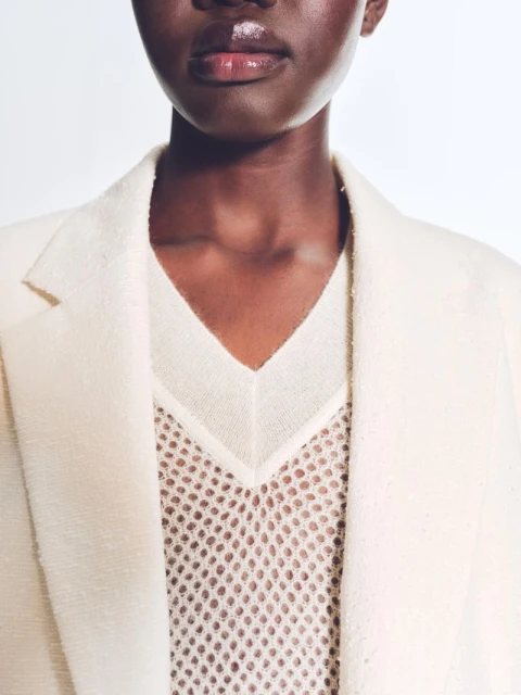A close-up studio shot features a Black woman wearing an off-white blazer and a mesh top, accentuated by glossy lips and neutral tones.