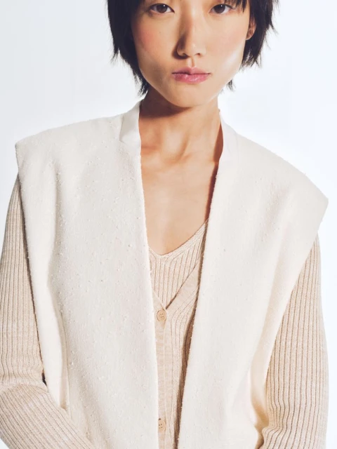 An Asian female poses in a studio wearing a cream-colored sweater and textured vest, captured in soft, diffused lighting against a white backdrop.