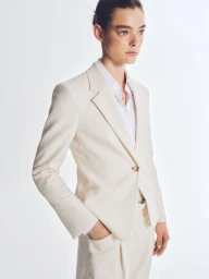Model wearing a cream blazer and white button-down shirt against a clean white seamless studio background.