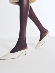 Close-up of legs in dark tights wearing white pointed-toe kitten heel mules against a clean white studio background.