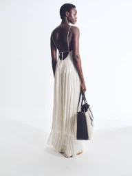 Model standing against white seamless background wearing a long white dress while holding a large tote bag.