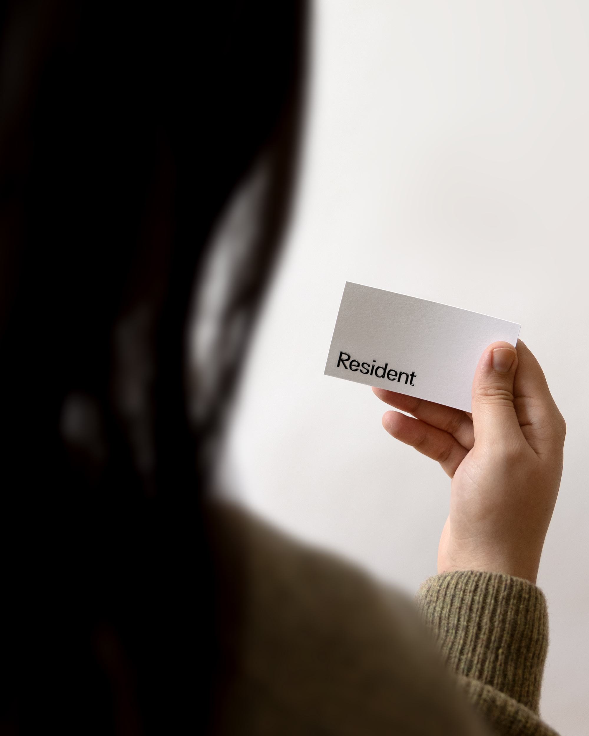 Unrecognizable person holds a white card with the word Resident printed in black letters, viewed from behind, against a plain background.