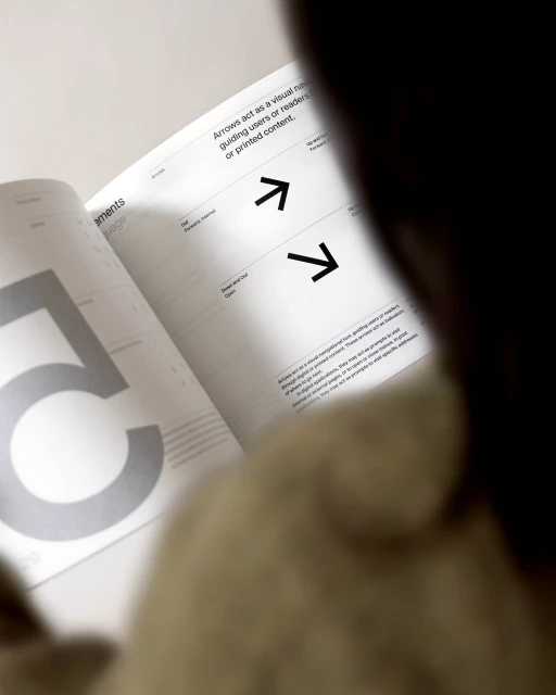 Open book with arrows pointing to written text; a person looks on. Book provides visual navigation for guiding users or readers.
