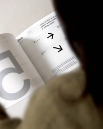 Over-the-shoulder view of a person reading an open brand guidelines booklet featuring typography and graphic arrow icons.
