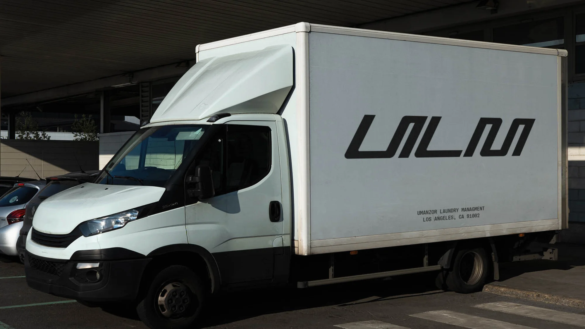 White delivery truck with a black logo and lettering sits parked on a city street in front of a building, with a parking lot nearby.