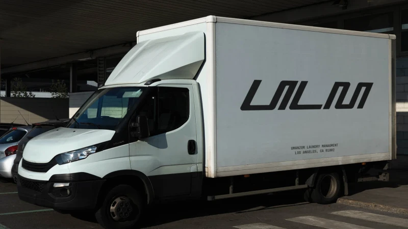 White delivery truck with a black logo and lettering sits parked on a city street in front of a building, with a parking lot nearby.