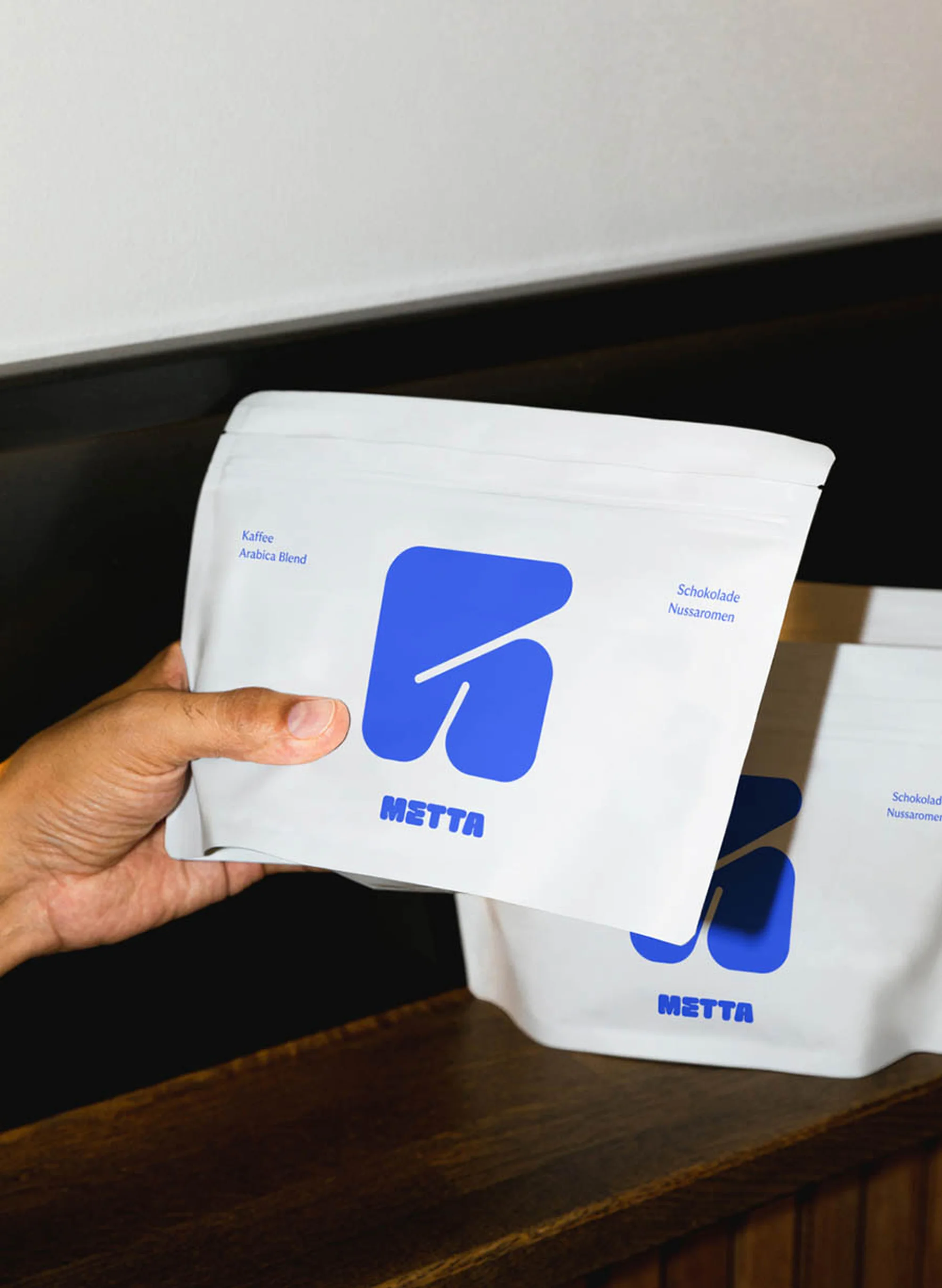 A hand holds a package of METTA coffee with a blue logo; another package sits on a wood surface against a black and white background.