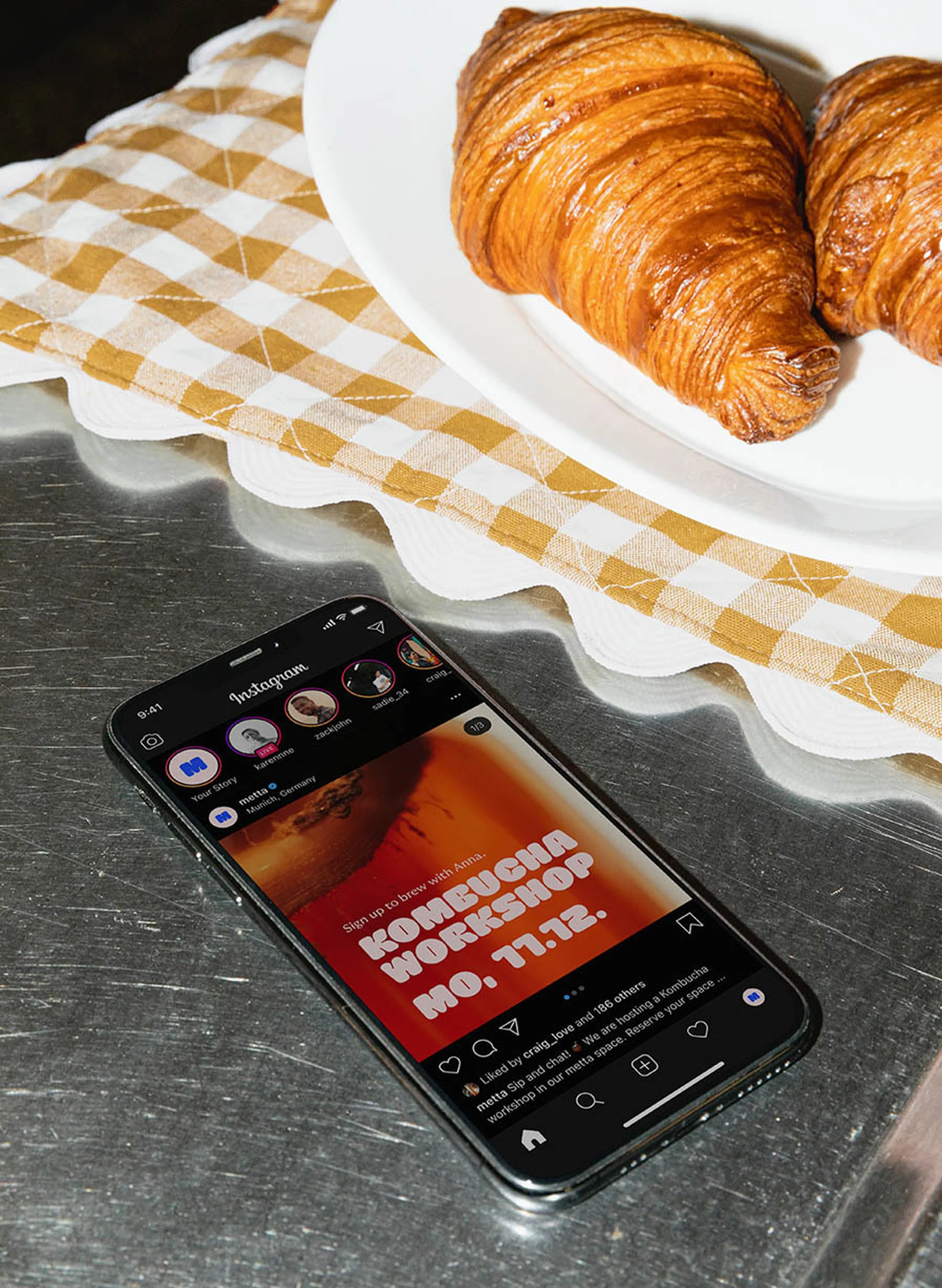 Croissants on a plate next to a smartphone displaying an Instagram post about a kombucha workshop, set against a checkered cloth.