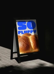 A-frame sidewalk sign displaying a poster with golden baked bread and bold blue text in a dimly lit setting.