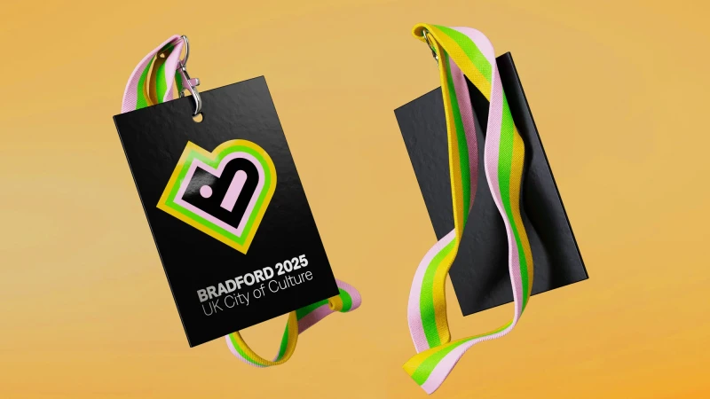 Two floating black event badges with multicolored striped lanyards, promoting Bradford 2025 UK City of Culture against yellow background.