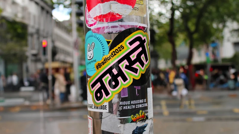 Close-up of a sticker-covered utility pole in an urban environment featuring stickers, including one with the Hindi word 'Namaste'.