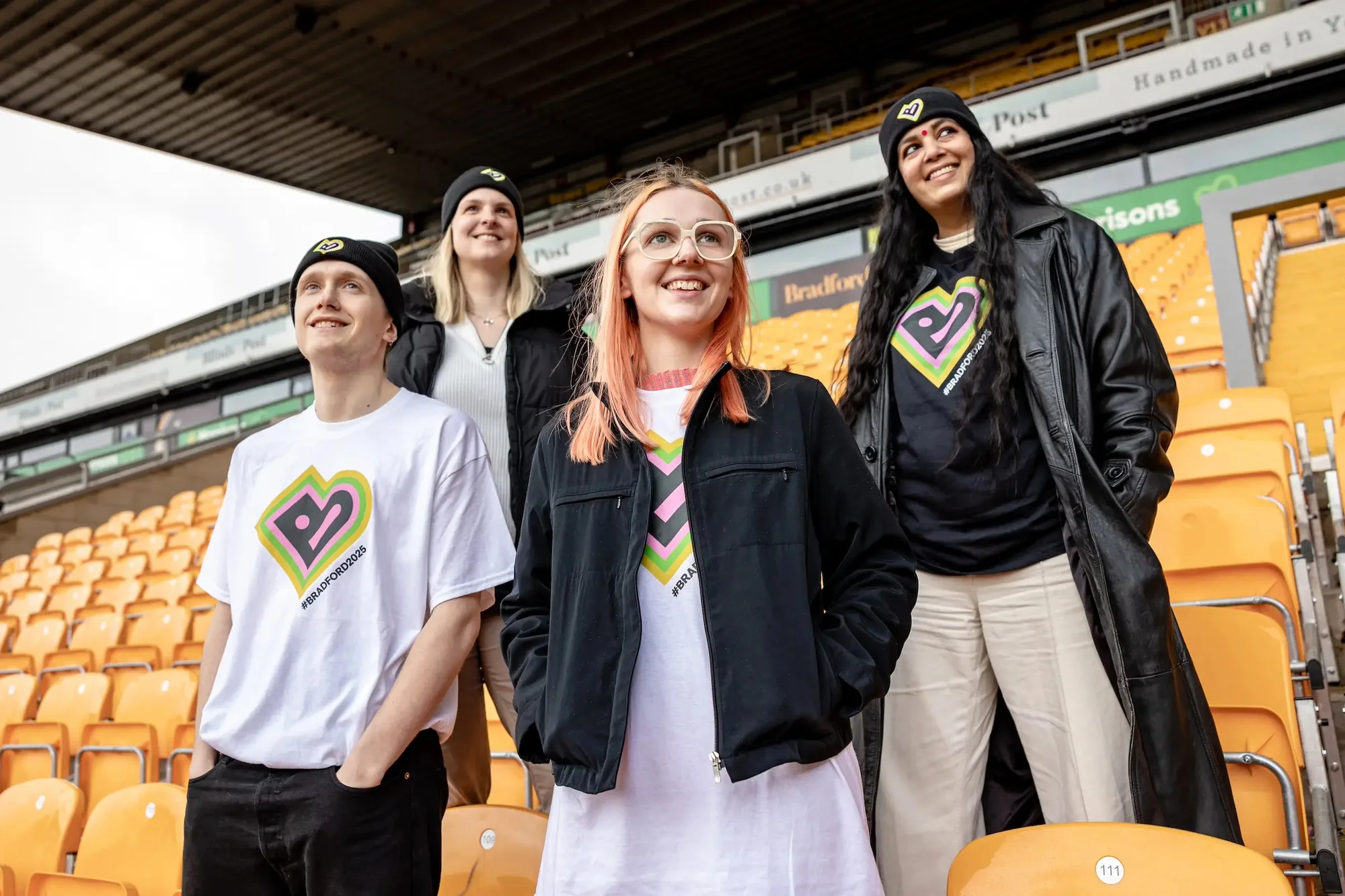 Group of young adults wearing branded T-shirts and hats at an outdoor stadium stand, smiling and looking upwards towards the sky.