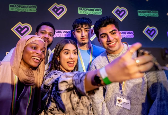A group of five smiling young adults pose for a selfie, showcasing a diverse group against a branded backdrop for 'Bradford 2025 UK City of Culture'.
