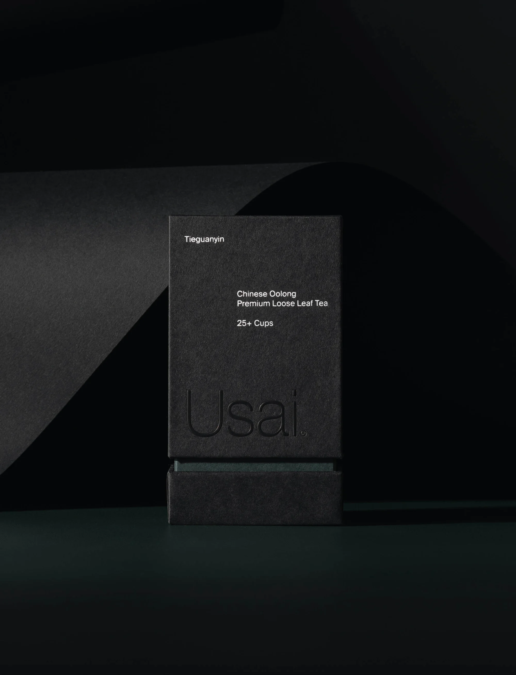 Elegant product shot of a black tea box with embossed branding on a dark green surface, set against a rippled black background.