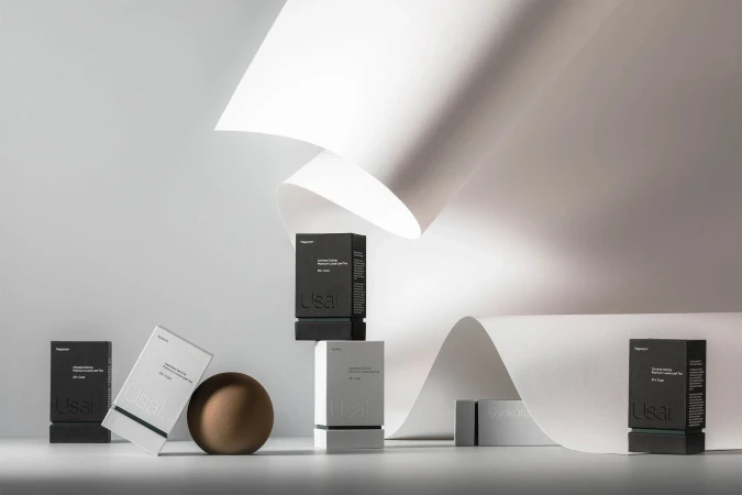Product photography of black and white boxes with the word Usal, arranged with geometric paper and a brown sphere in a studio.