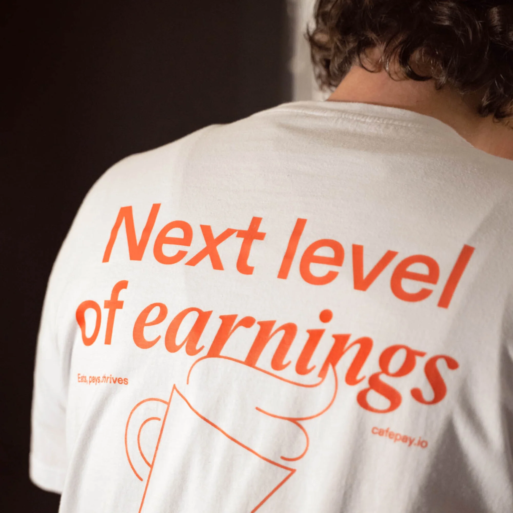 Back of a white t-shirt with red text 'Next level of earnings,' a stylized cup outline, and cafepay.io logo, set against a dark background.