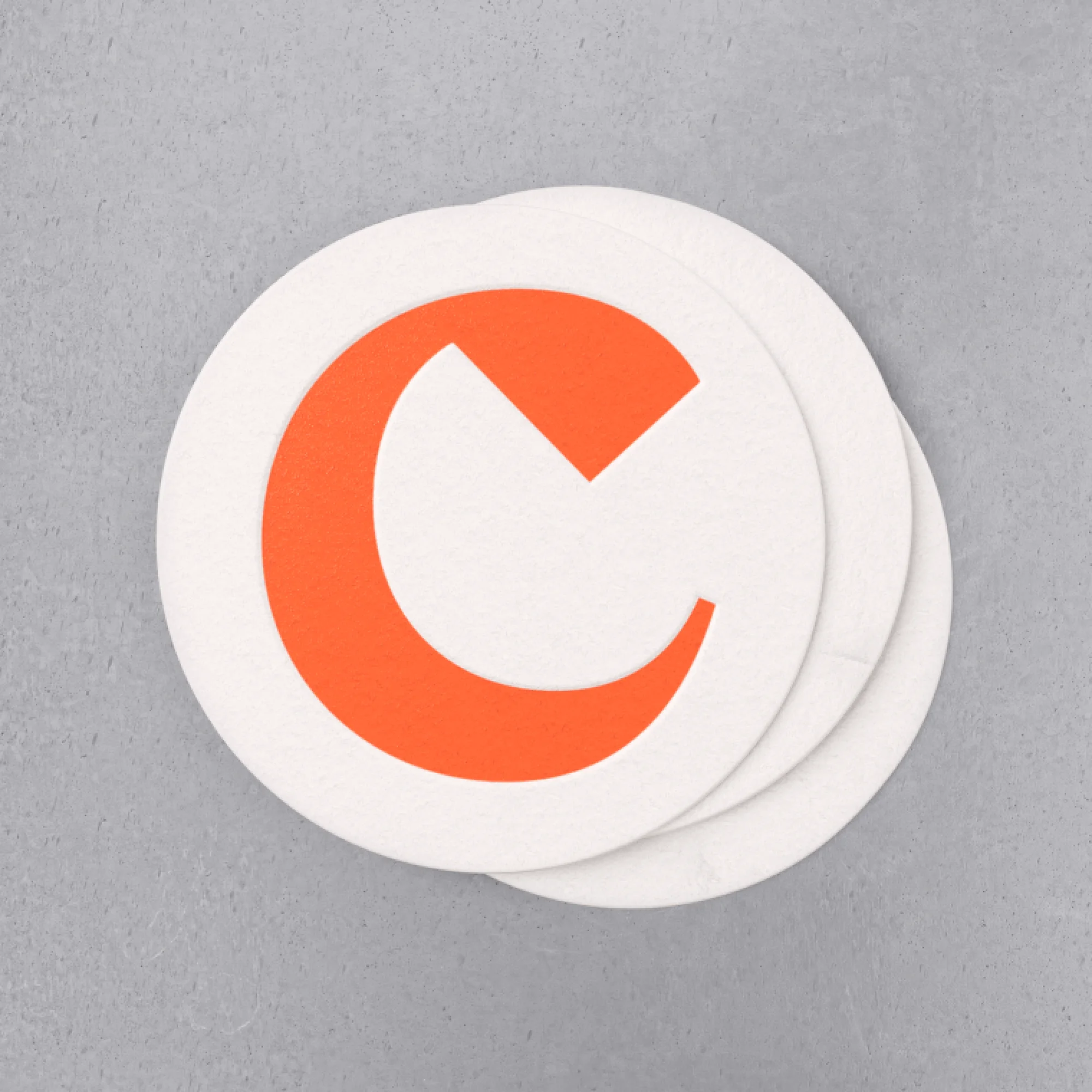 Close-up of a stack of three white round coasters, each with a large, vibrant orange letter C, placed on a textured gray surface.