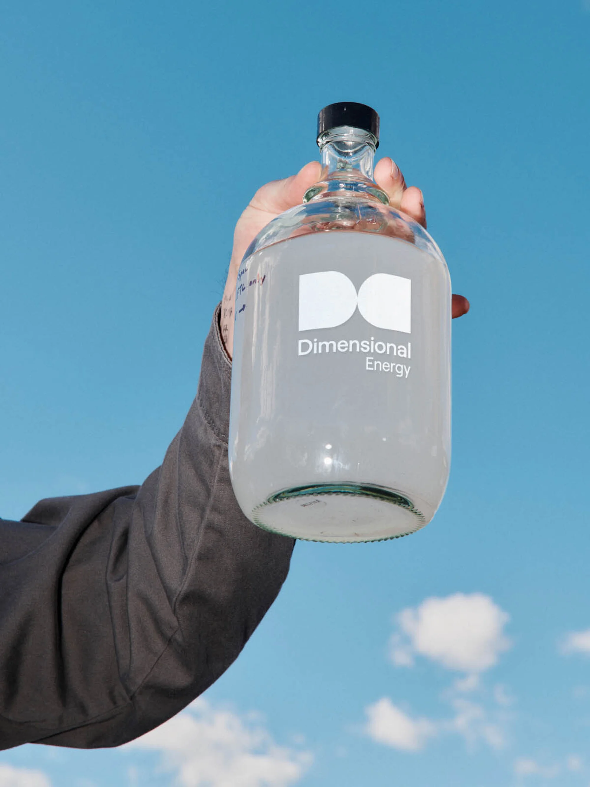 Hand holding a clear glass jug labeled 'Dimensional Energy' against a blue sky with clouds in a bright, sunny, outdoor setting.