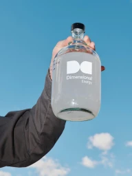 Hand holding up a clear glass bottle with a white logo against a bright blue sky with white clouds