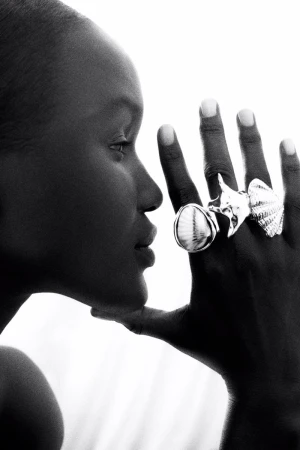 Close-up black and white portrait of a Black woman with statement shell rings on her hand, set against a bright white background.