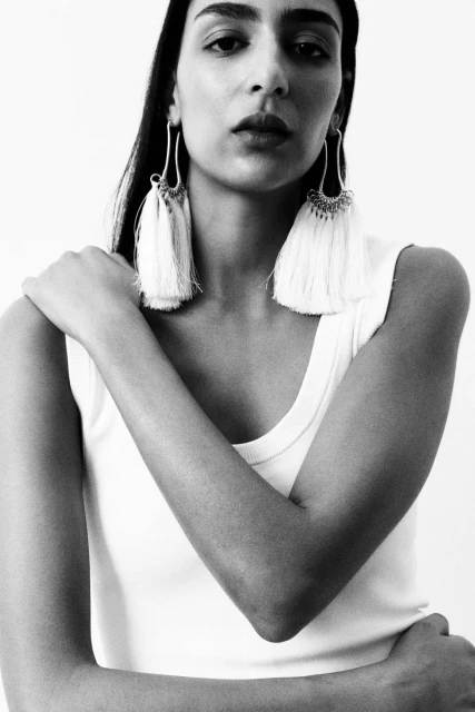 Black and white portrait of a woman with dark hair wearing a white tank top and tassel earrings against a white background.