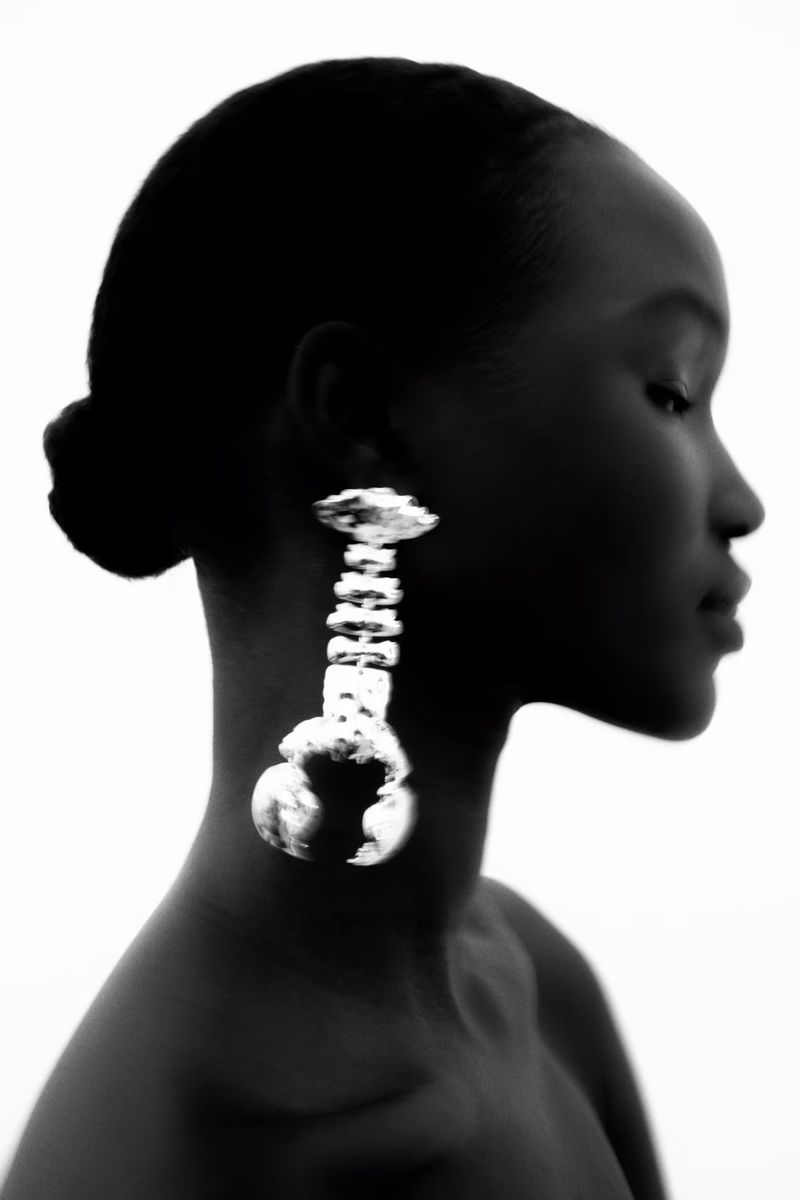 Portrait of a Black woman in profile wearing a large silver statement earring against a white background in a photo studio.