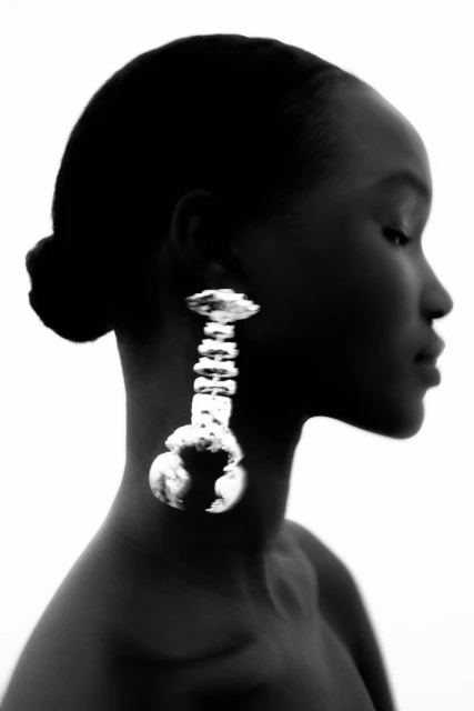 Portrait of a Black woman in profile wearing a large silver statement earring against a white background in a photo studio.