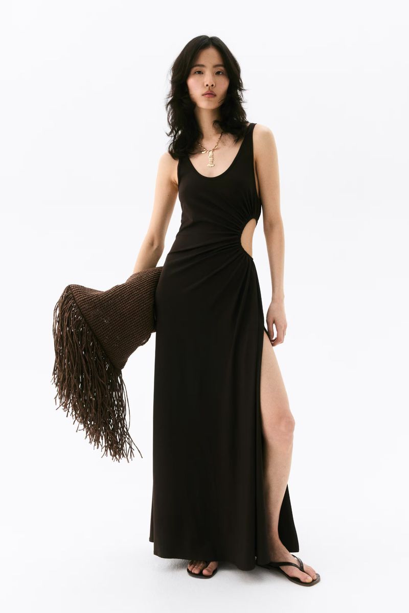 A fashionable Asian woman poses in a black dress with a high slit, holding a brown, fringed shawl against a bright white backdrop.