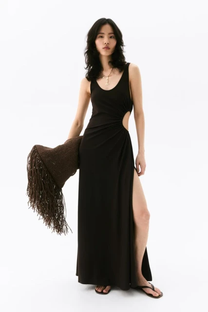 A fashionable Asian woman poses in a black dress with a high slit, holding a brown, fringed shawl against a bright white backdrop.